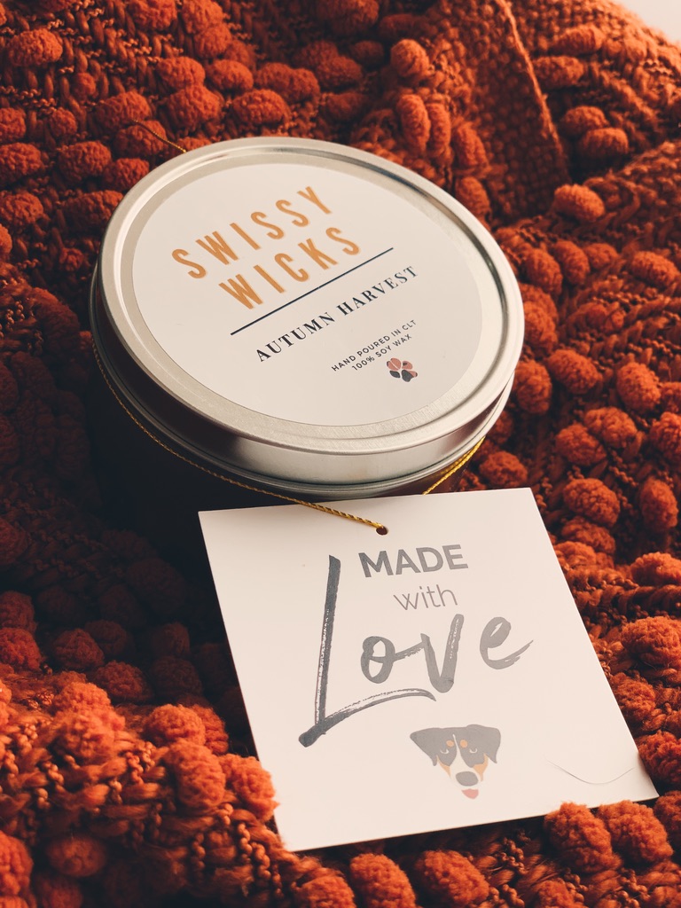 A Swissy Wicks candle tin with an Autumn Harvest label and a 'Made with Love' tag showing a dog's face